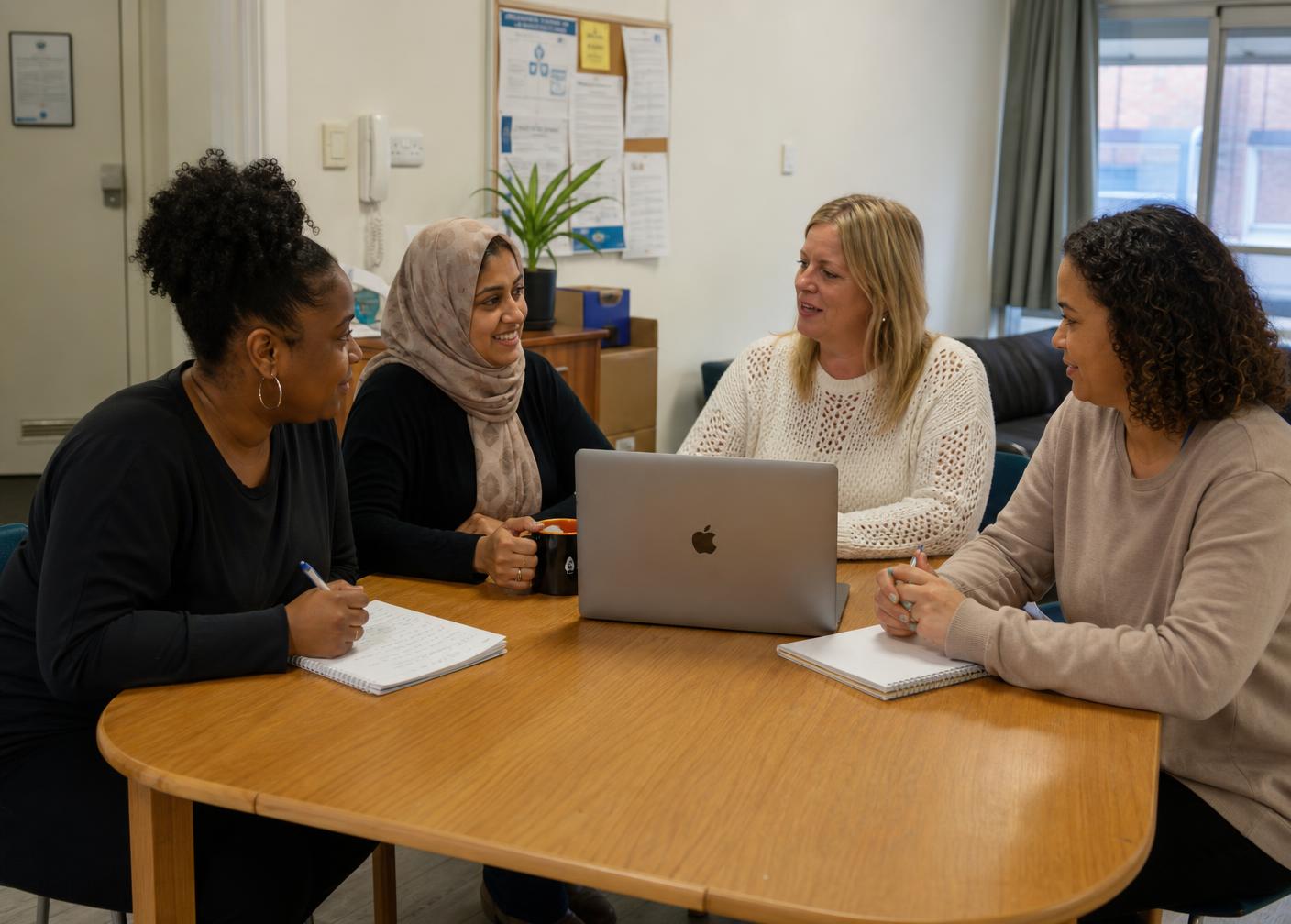 A diverse team of four women support workers talking together around a table with notebooks and a laptop