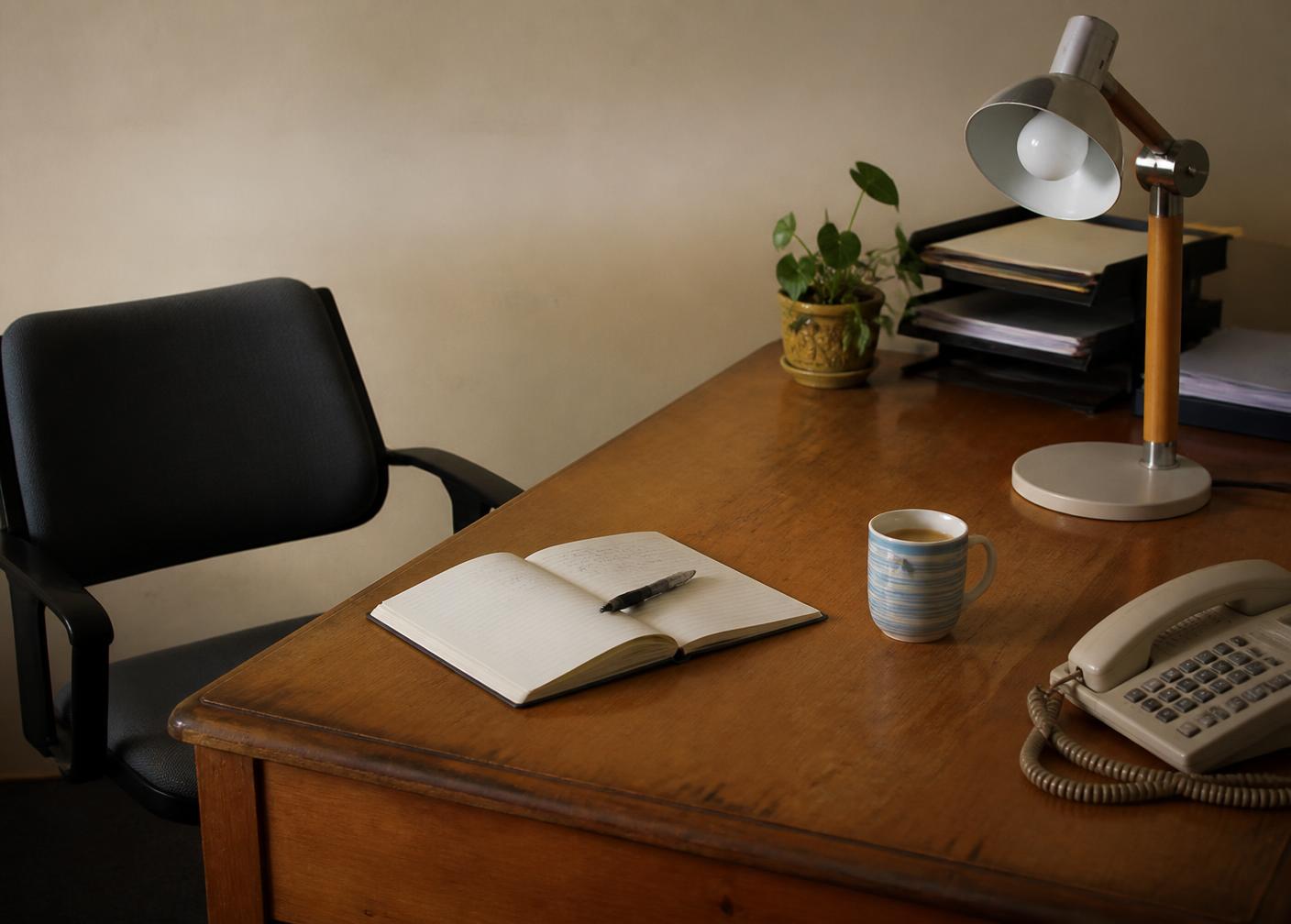 A simple, welcoming office desk with an open notebook, pen, telephone, lamp and a mug of tea