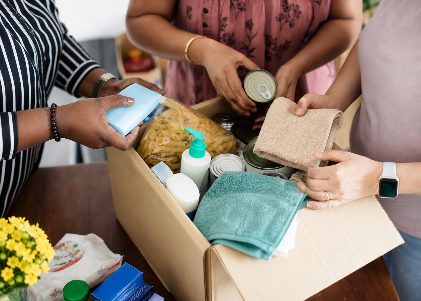 Three women volunteers of different backgrounds packing toiletries, towels, food staples and a children's book into a box and tote bag