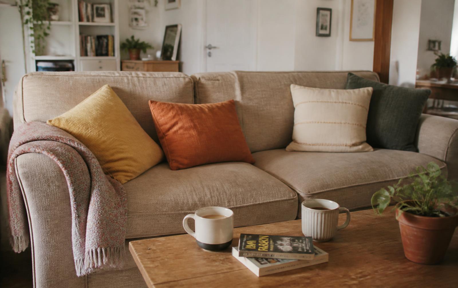 A warm, homely living room with a comfortable sofa, cushions, a throw, books and two mugs of tea on a simple wooden table