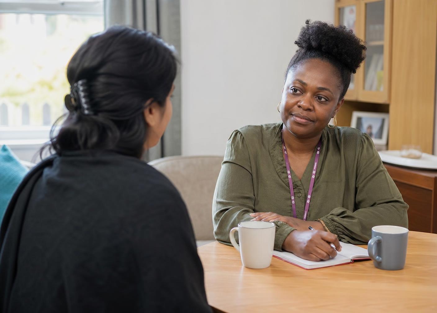 A female support worker listening attentively to a woman during a calm, confidential conversation over notebooks and tea