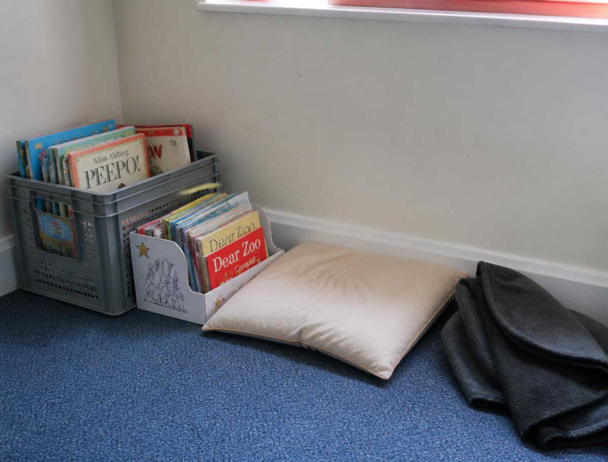 A small reading corner with a crate of children's picture books, a cushion and a folded blanket