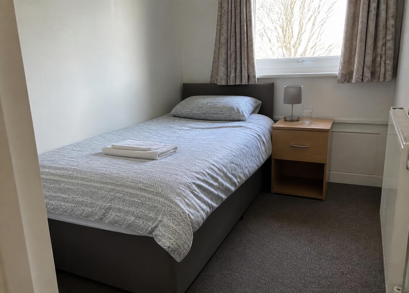 A simple, clean bedroom with plain bedding, folded towels and a practical bedside table with a lamp